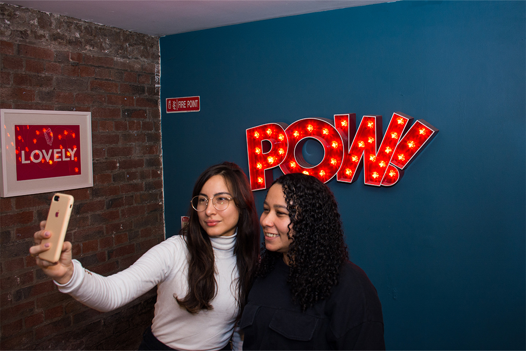 Two students taking a selfie beside a bright “POW” sign inside ISI Dublin.